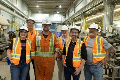•	A group of people wearing safety vests and helmets stand in front of machinery at pulp and paper mill.