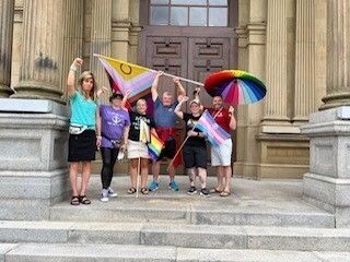 A group of six people standing on steps outside waving trans and pride flags