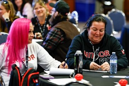 two women sitting at a table smiling, others at tables in the background