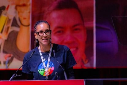 A black women wearing glasses and a lanyard speaks at a podium.