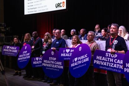 People standing in front of a stage carrying purple signs that read, “Stop health care privatization,”
