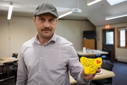 A man in a hat holds up a yellow rock that reads, love yourself