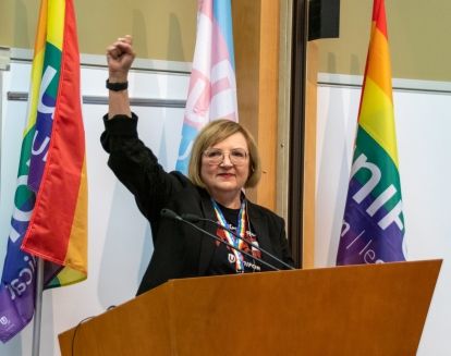 Lana Payne holding up a fist standing behind a poidum in front of two pride flags on either side.