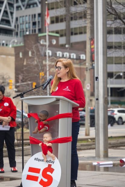 A women speaking at an outdoor podium, an equal pay day sign leaning on the podium.