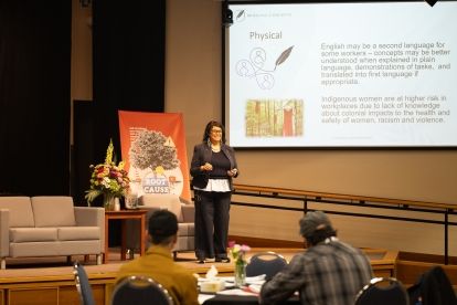A women speaking on stage with a screen, x-stand and couch in the background