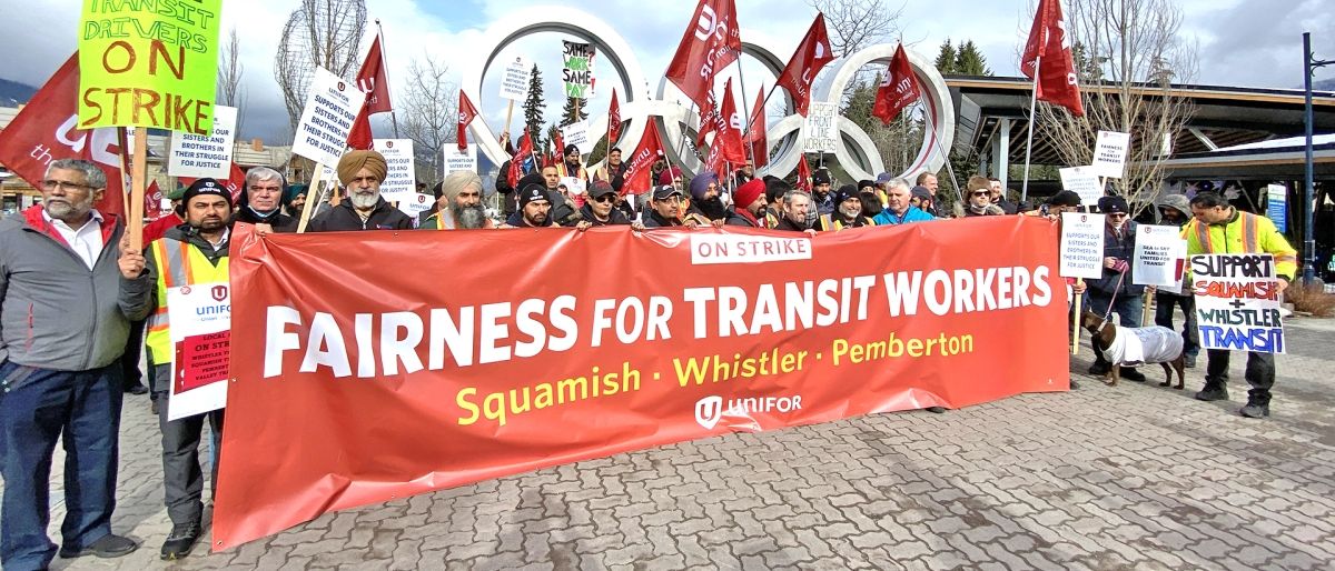 Large group of rally participants with flags and placards posing with a "Fairness for Transit Workers" banner in front of Whistler Village's Olympic rings..