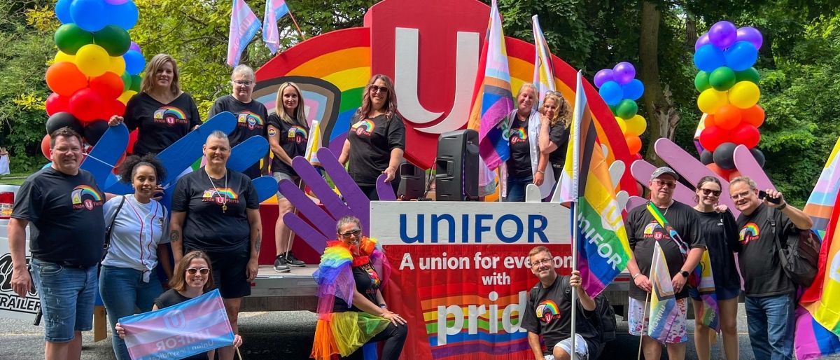A large group of Unifor members standing in front of a painted rainbow with a Unifor shield.