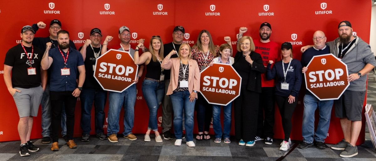 A large group posing in front of a Unifor banner with three holding stop signs