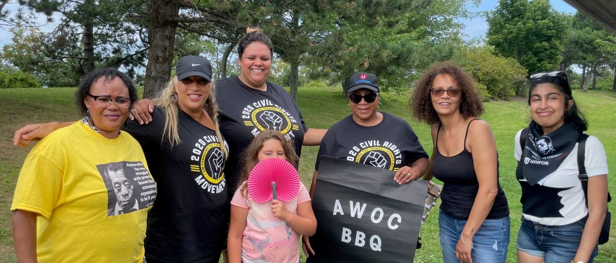 Group of people standing together with tree in the background, one person near the middle holding black sign that says, “AWOC BBQ.”