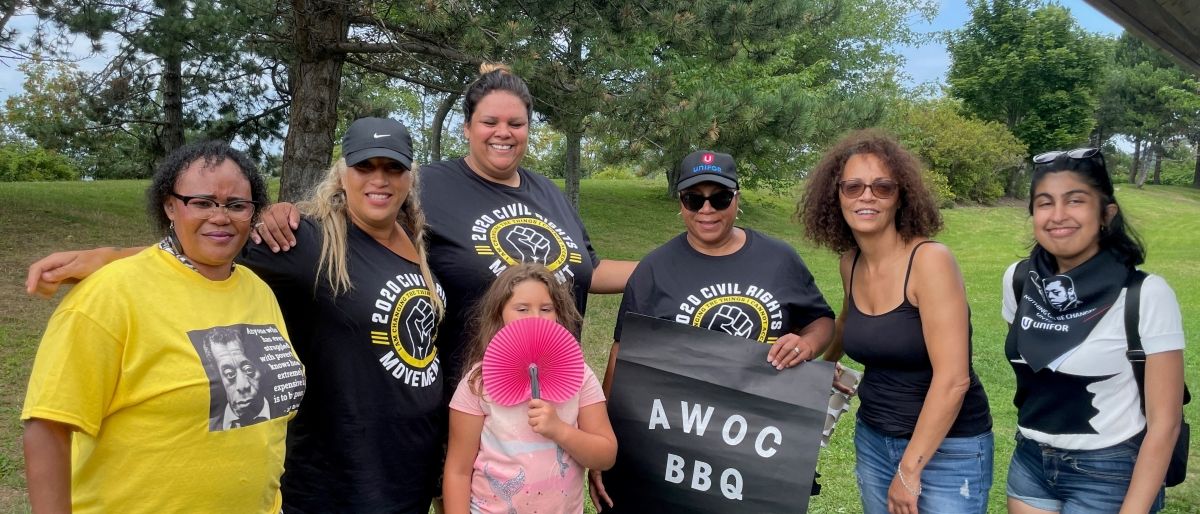 Group of people standing together with tree in the background, one person near the middle holding black sign that says, “AWOC BBQ.”