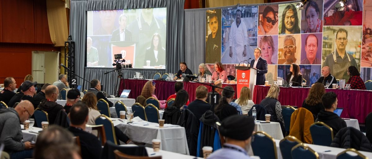 A large room with people seated classroom style and a person speaking from a podium.