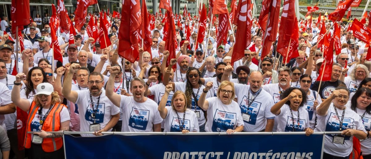 A huge rally of people standing behind a Unifor "Protect Canadian Job" banner holding red Unifor flags