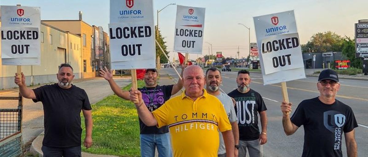 Six men stand on a sidewalk holding signs that read "UNIFOR LOCKED OUT," indicating a labor dispute.