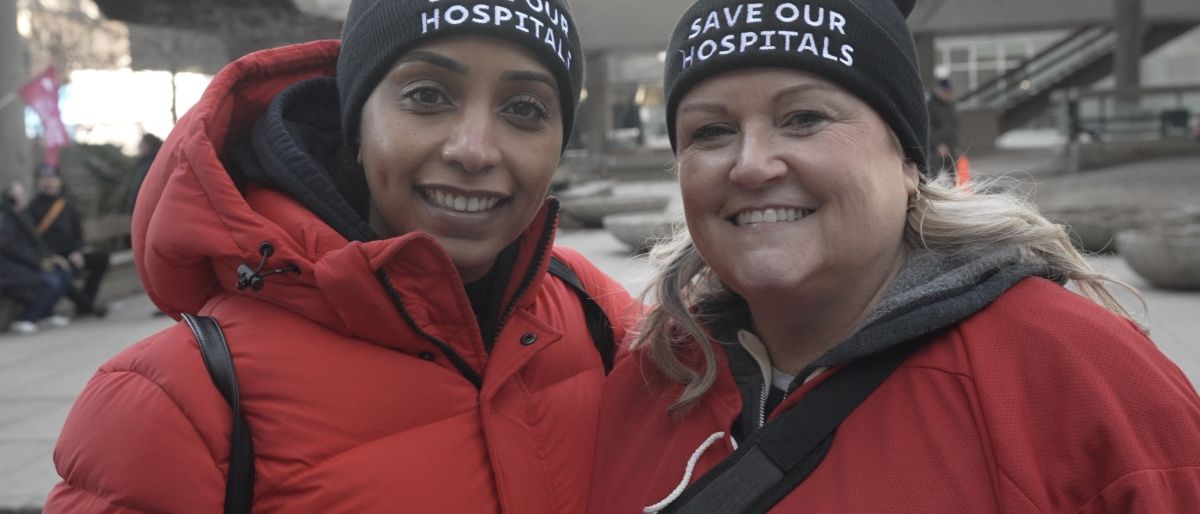 Two Unifor members wearing red jackets with black hats that say "Save our Hospitals"