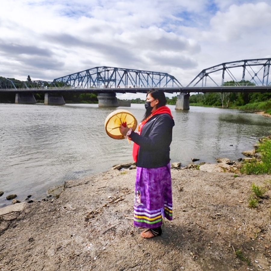 Woman standing on riverbank holding a drum. A bridge is visible in the background.