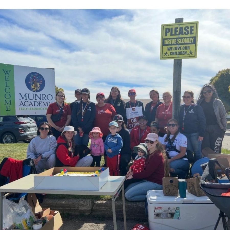  A group of women and three children, some wearing red and holding "On Strike" signs, gather outside near Munro Academy banners and a "Please Drive Slowly, we love our children" sign on a sunny day, at a picket line. 