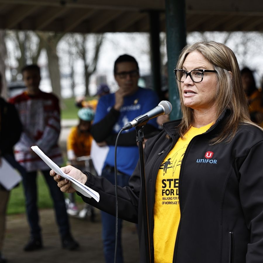 Joanne Hay stands at a microphone, speaking to gathered crowd