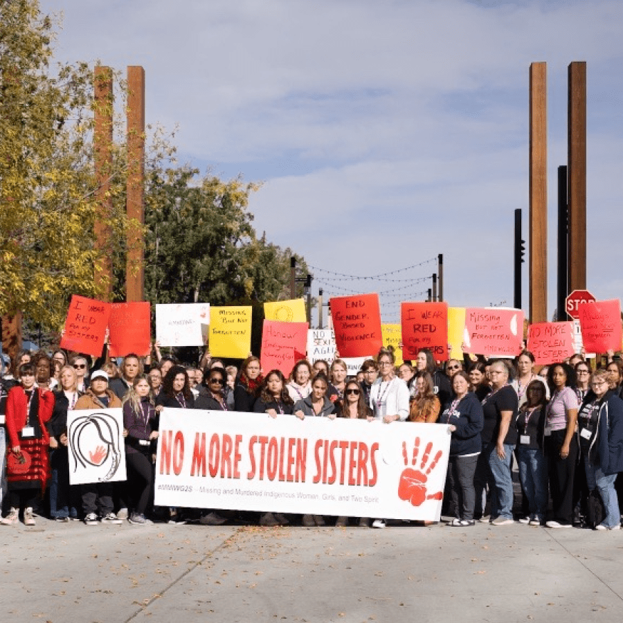 A large group of women hold a banner "No more stolen sisters" and coloured placcards up high